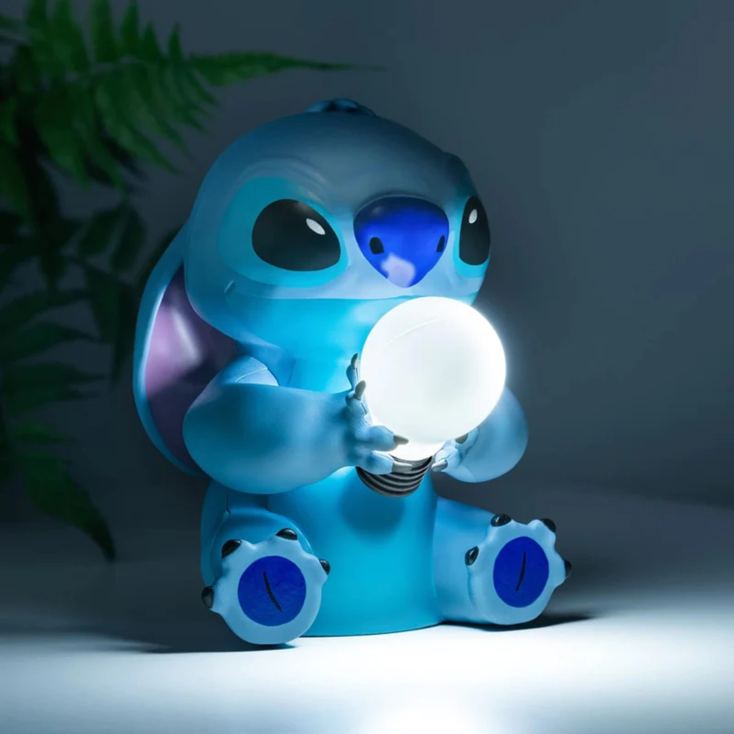 Een blauw Stitch-figuur houdt een gloeiende gloeilamp vast. Donkere kamer met groene plant op de achtergrond.