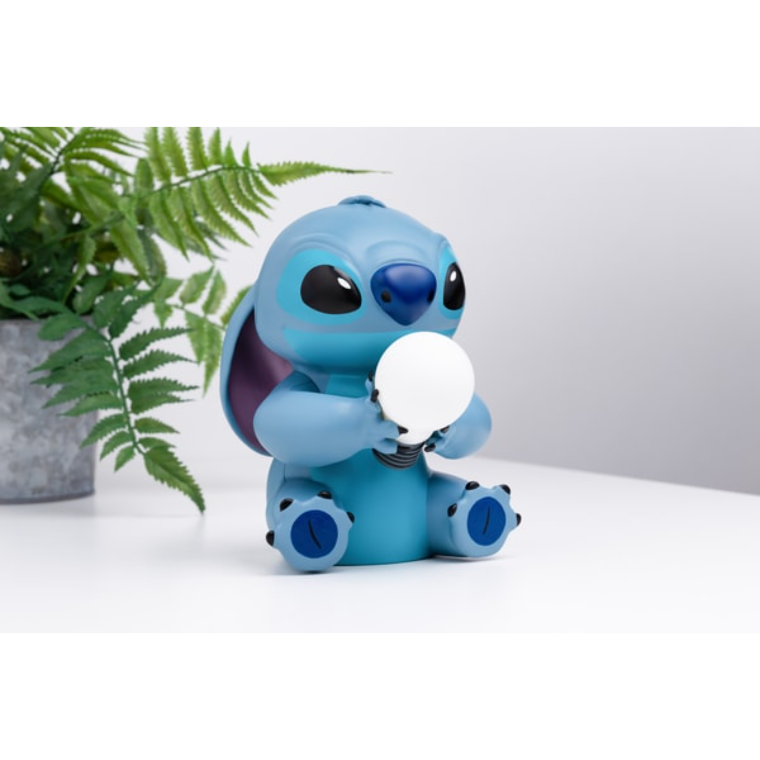 Stitch-beeldje met een witte gloeilamp, met groene varen en een witte achtergrond.