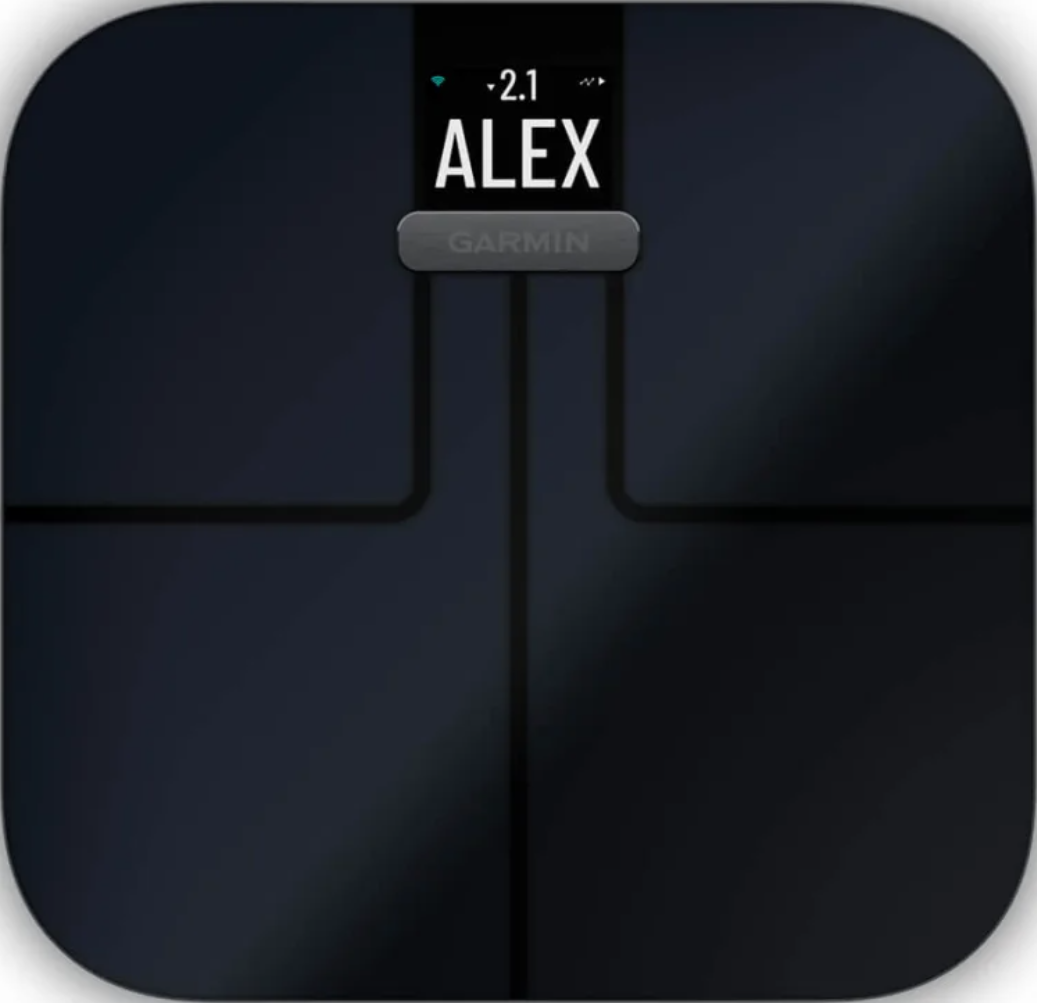 Bilancia intelligente Garmin nera che mostra 'ALEX' e un peso di 2.1. Dispositivo scuro e quadrato.