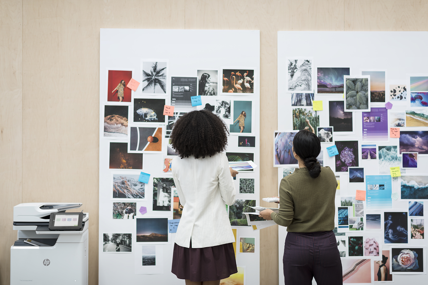 Two women looking at a display of pictures on a wall with an HP printer on the left.