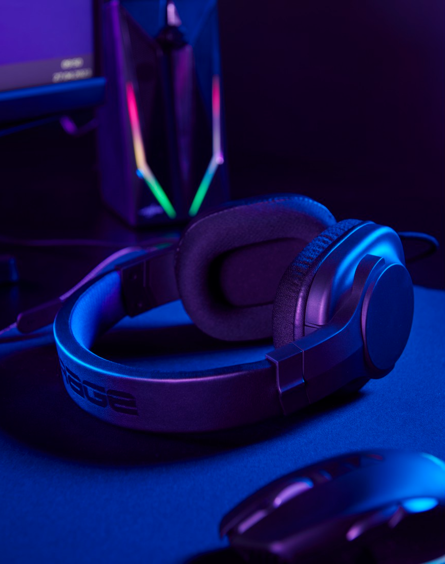 Fekete gamer fejhallgató sötét felületen, monitorral a háttérben. Neonfények gamer hangulatot keltenek.