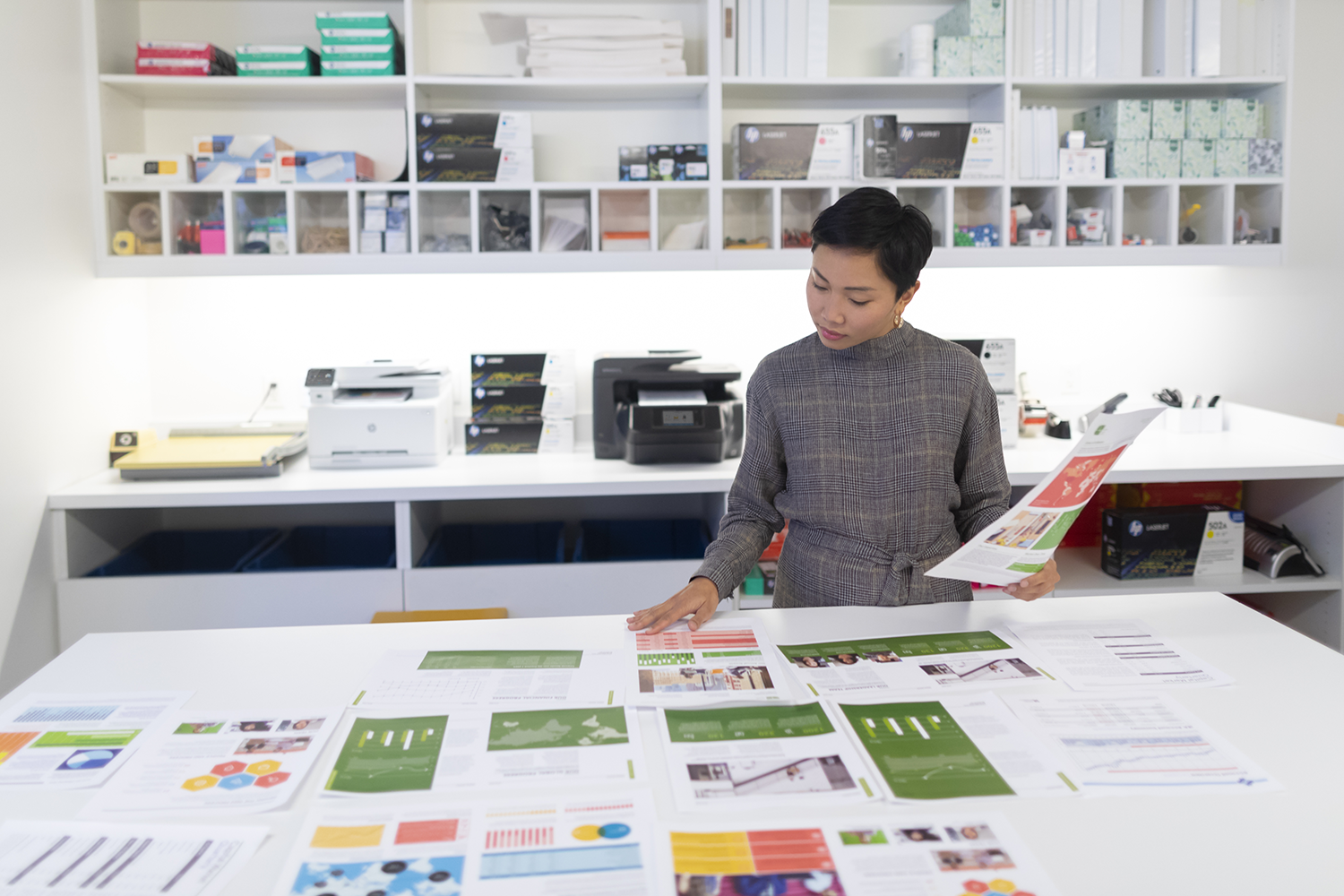 A woman reviewing printouts on a white table with various documents and printers.