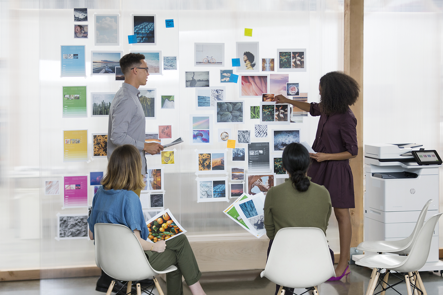 Four people around a clear wall with pictures, with a printer and chairs.