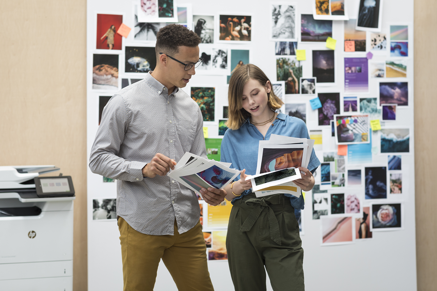 Two people looking at printouts in front of a board with photos and notes.