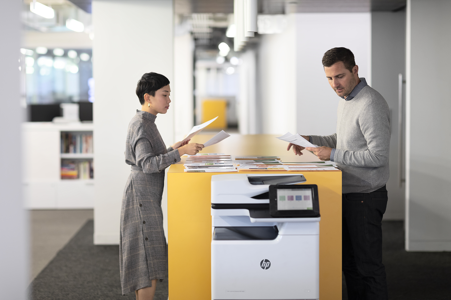 Two people looking at documents next to a yellow printer. The background is a bright office.
