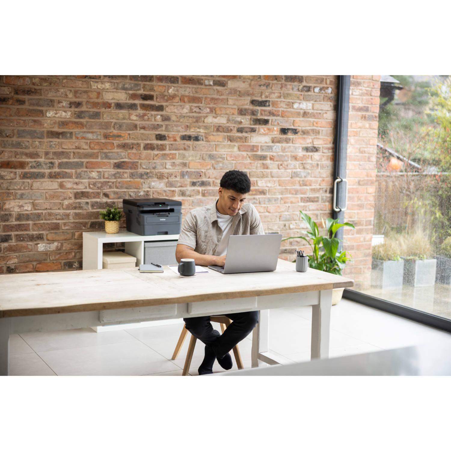 Man works on a laptop at a wooden desk. A printer, mug, and pens are nearby. Brick wall and window are in the background.