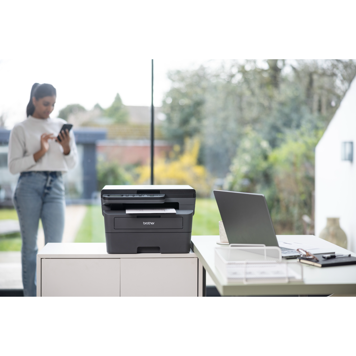 A woman using a phone stands near a printer on a white table.