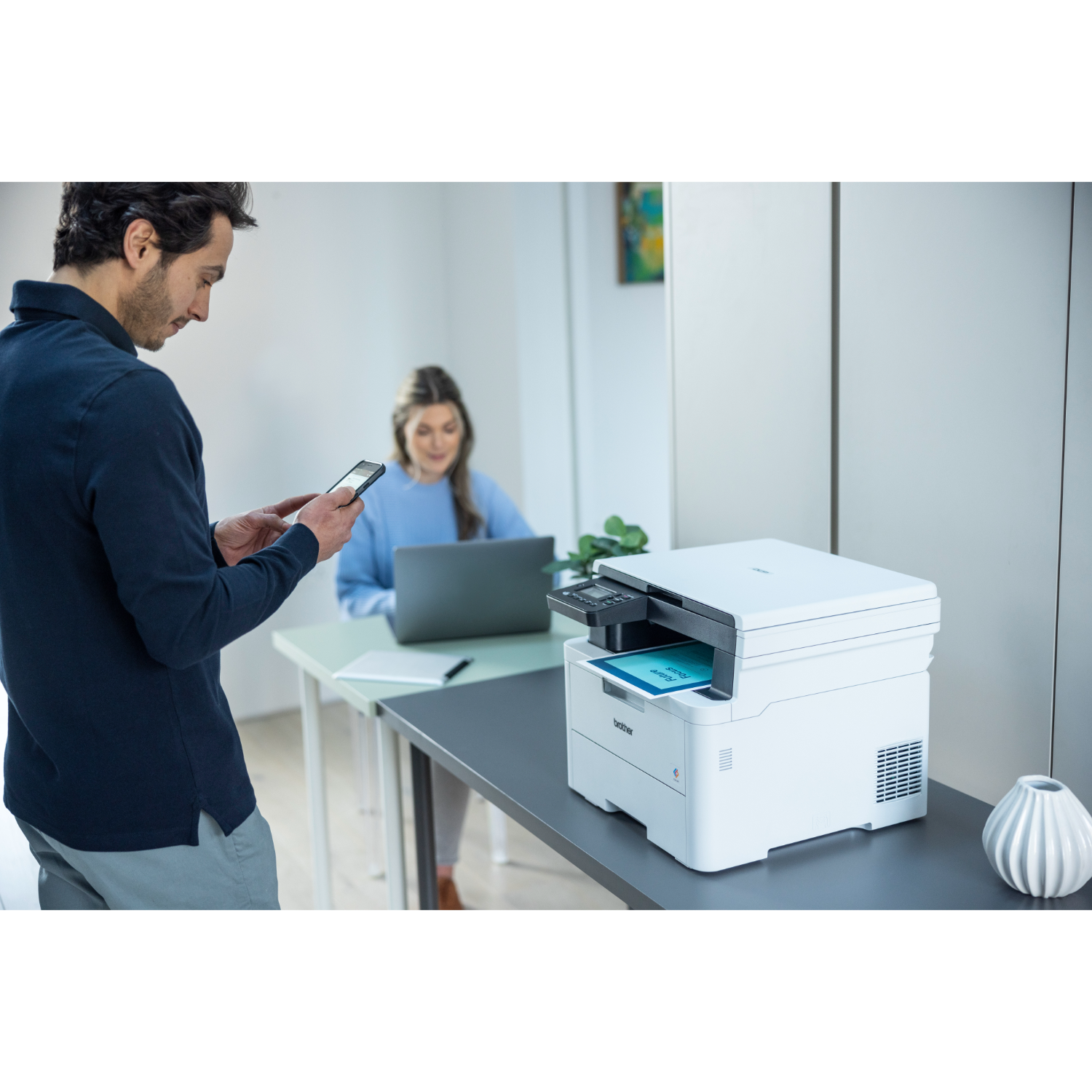 Man using phone, woman at laptop, printer on desk. Office setup with white walls and gray desk.