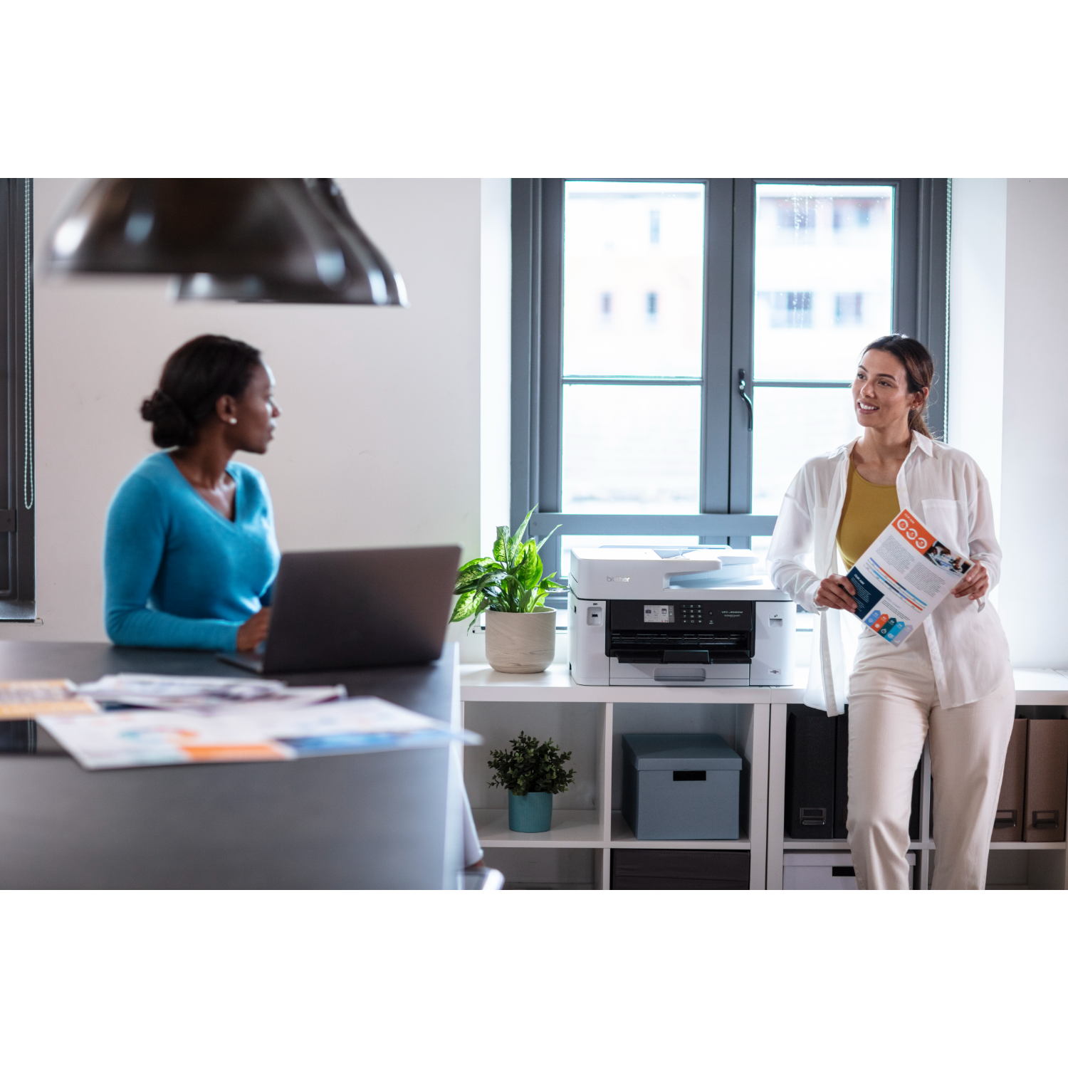 Two women in an office. One sits with a laptop, the other holds a brochure.