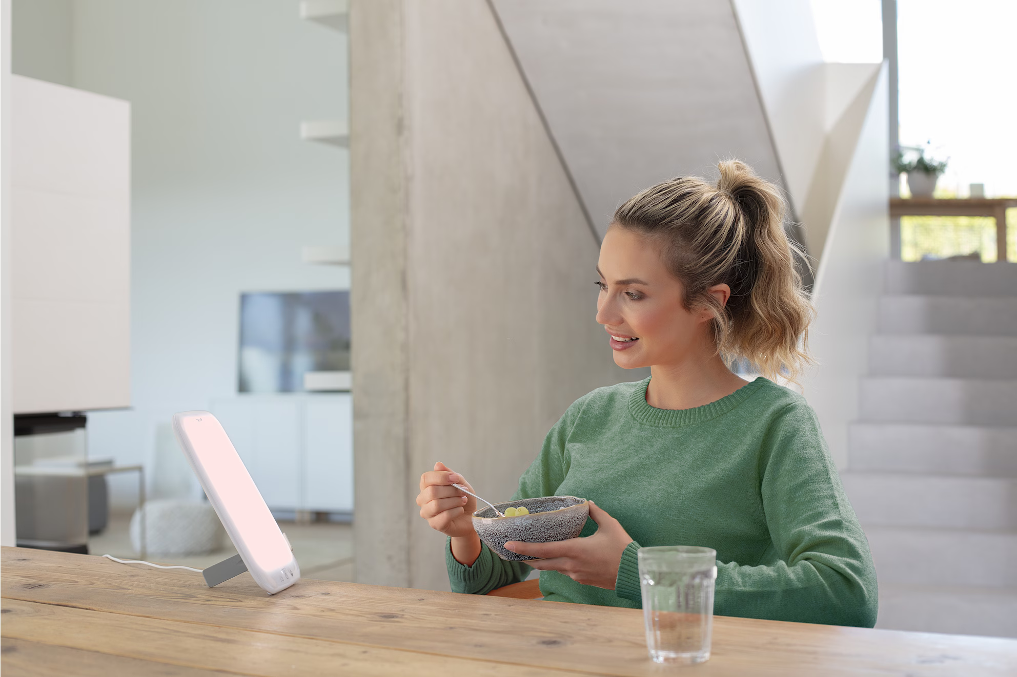 Femme mangeant dans un bol, assise à une table avec une lampe de luminothérapie. Pièce lumineuse.
