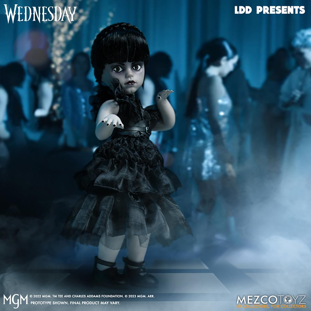 Wednesday Addams Puppe. Sie trägt ein schwarzes Kleid. Der Hintergrund hat verschwommene Figuren.
