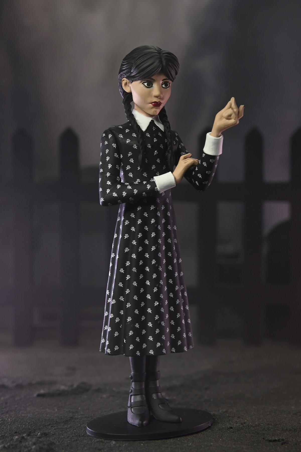 Wednesday Addams-Figur. Sie trägt ein schwarzes Kleid, Zöpfe und steht vor einem Zaun auf einem schwarzen Sockel.