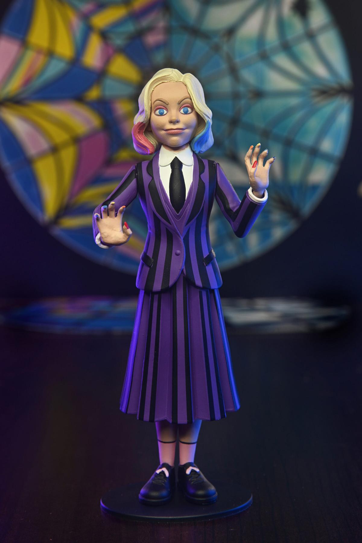 Eine Wednesday Addams-Figur in einem lila-schwarzen Anzug und Rock, stehend auf einem schwarzen Sockel.