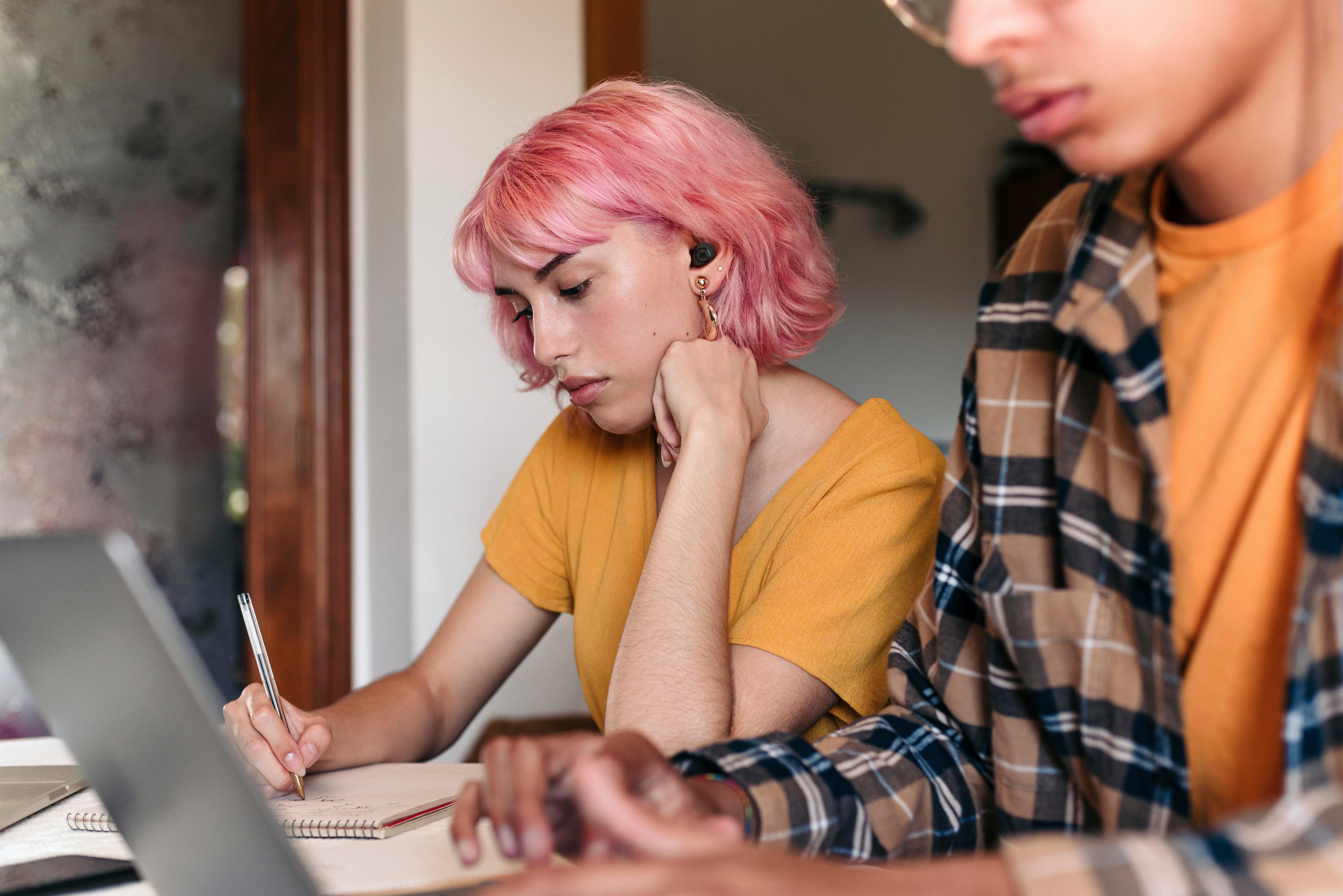 Eine Frau mit pinkfarbenen Haaren schreibt in ein Notizbuch. Eine andere Person benutzt einen Laptop neben ihr.