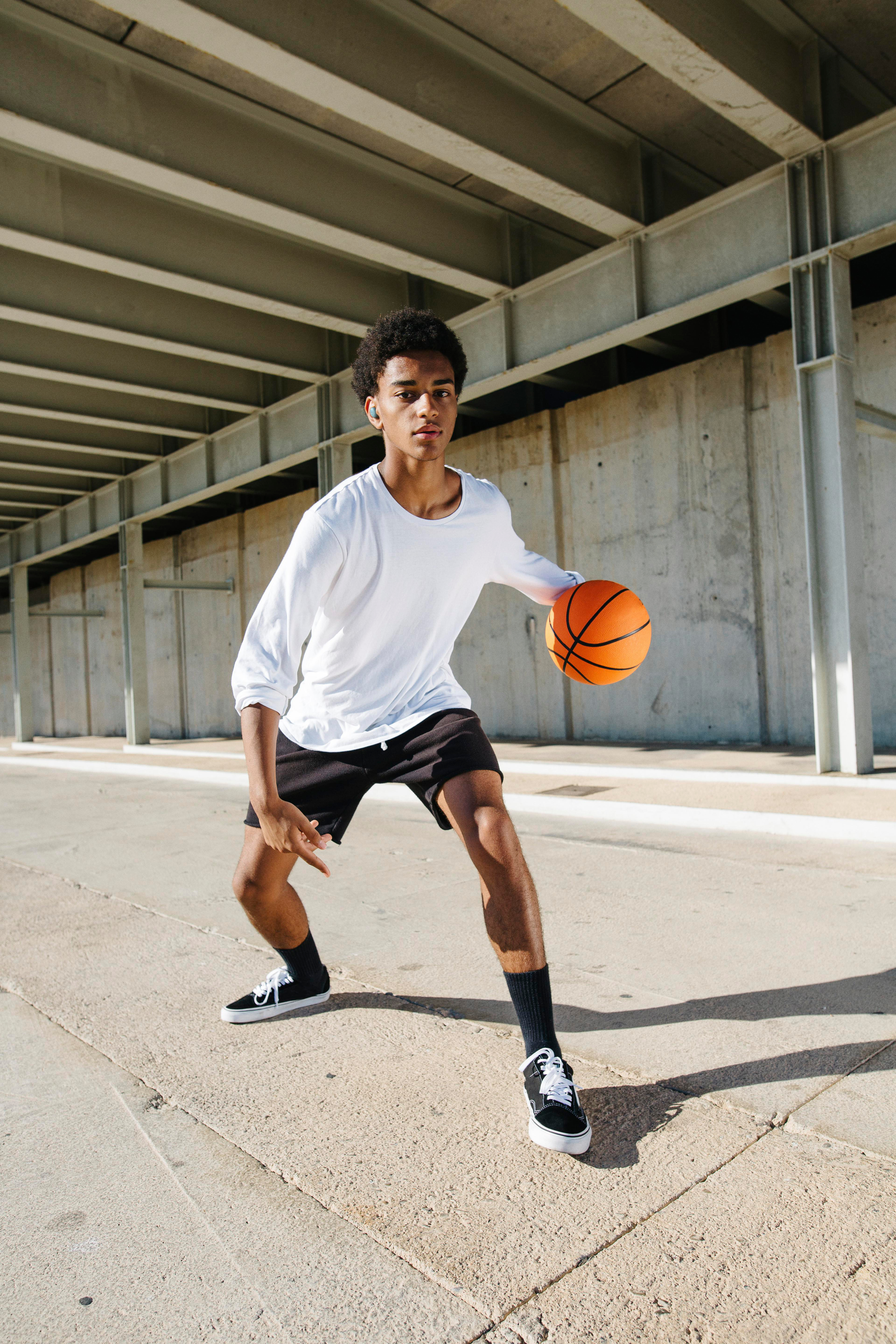 Ein junger Mann dribbelt einen Basketball unter einer Betonkonstruktion. Er trägt ein weißes Hemd und schwarze Shorts.