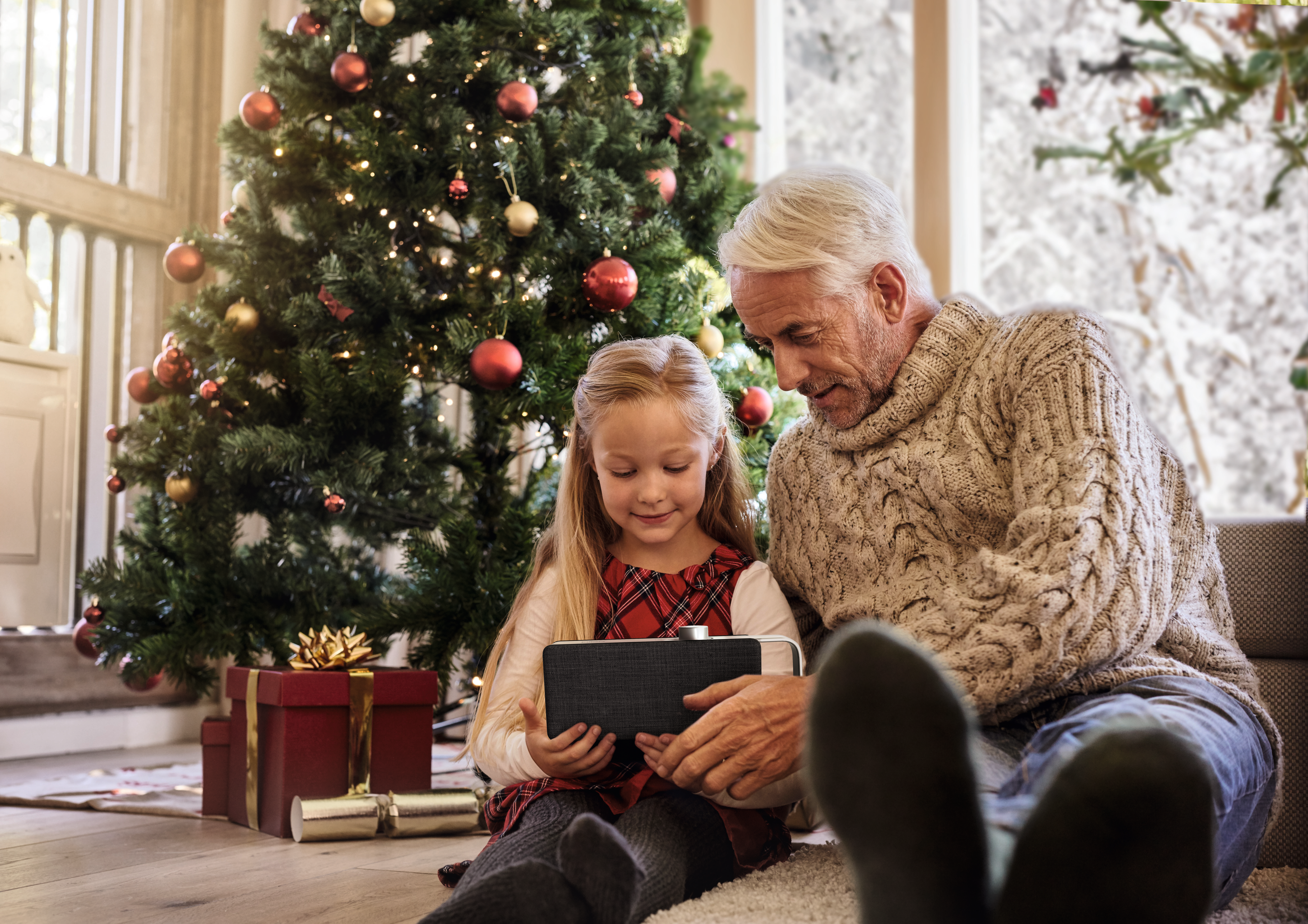 Una bambina e un uomo guardano un tablet vicino a un albero di Natale. Sono presenti dei regali.