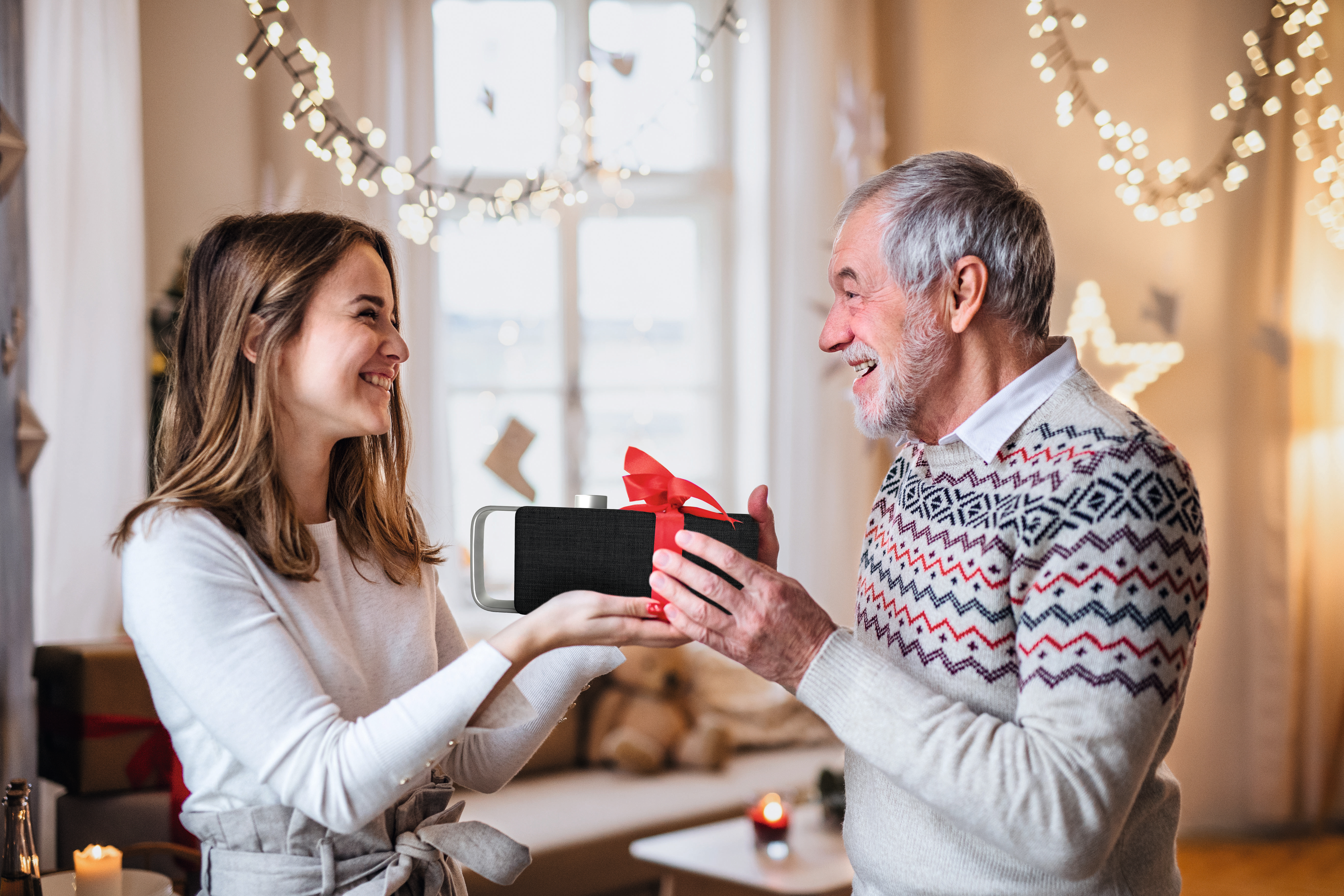 Una donna regala un regalo a un uomo anziano, sorridente, luci festive sullo sfondo.