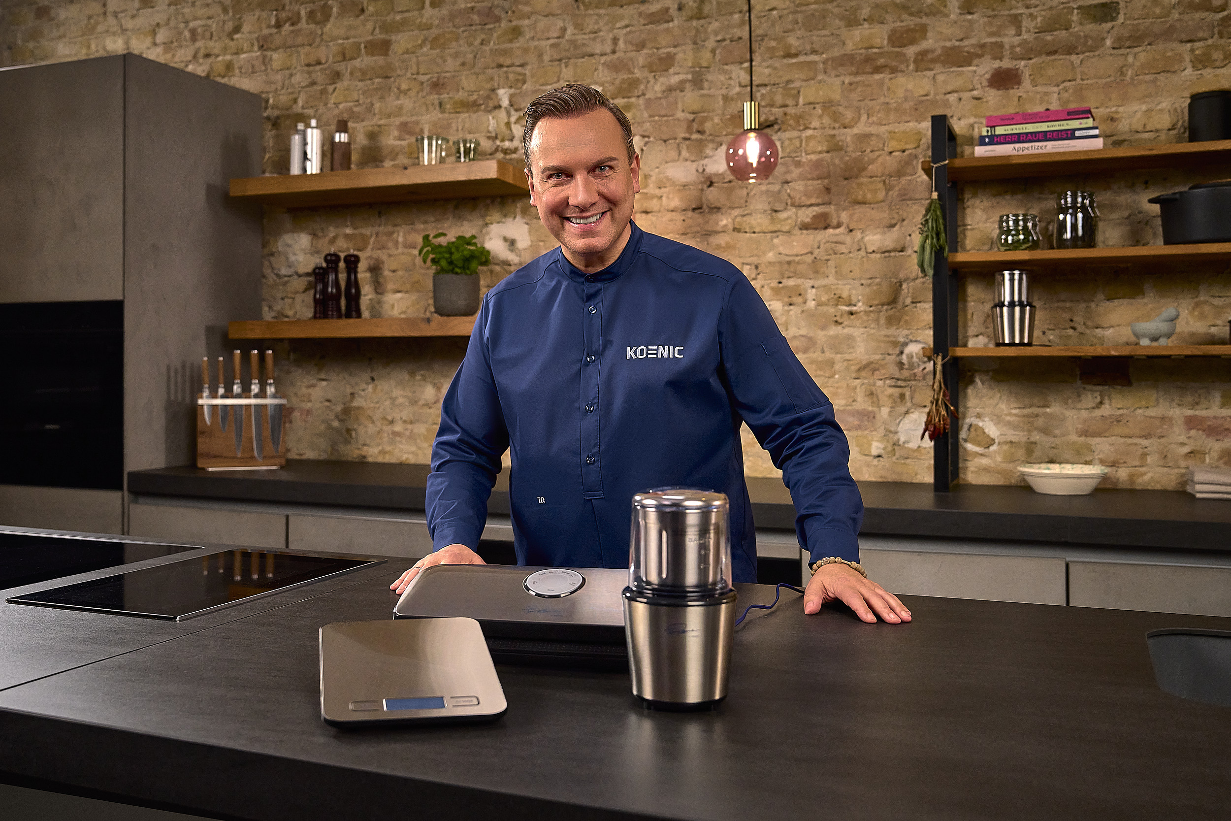 Mann in einer Küche, lächelnd, neben einer Waage, einer Kaffeemühle und einer Arbeitsplatte.