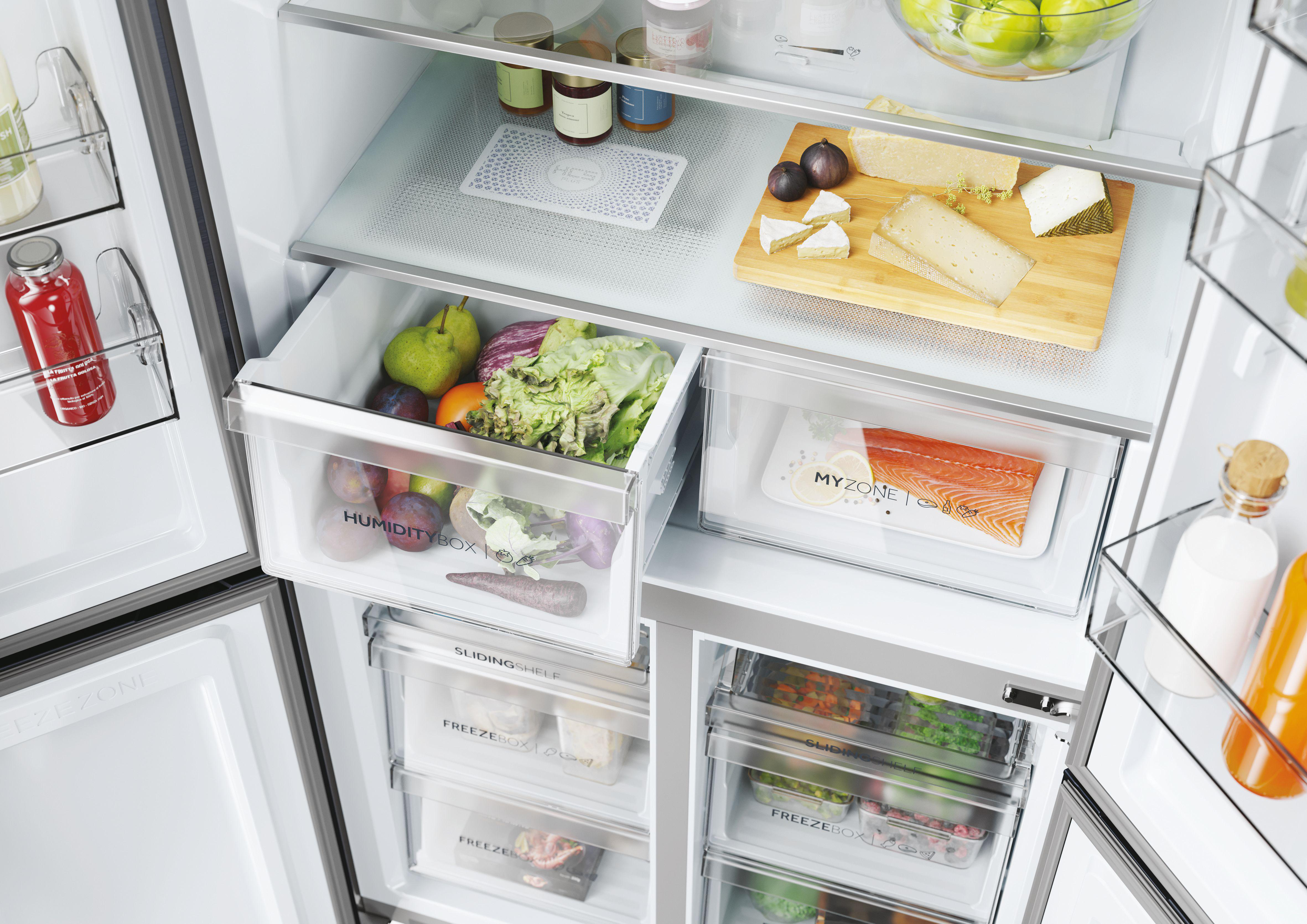 Offener Kühlschrank mit frischen Produkten und Lebensmitteln in Schubladen und Regalen. Früchte, Gemüse und verpackte Waren sind sichtbar.