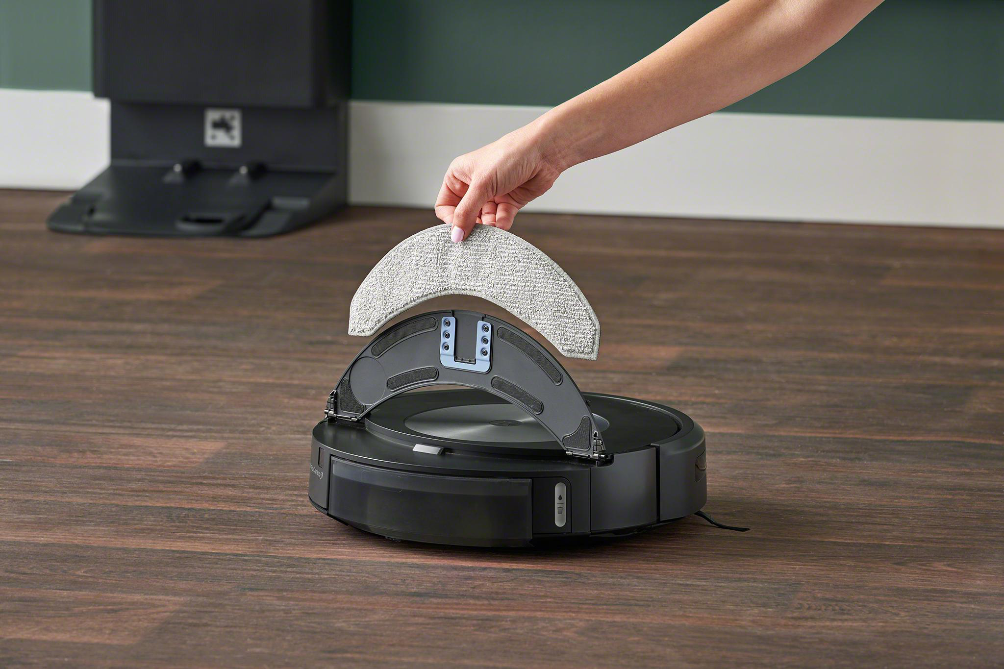 Eine Hand hält ein Reinigungspad über einem schwarzen Roboter-Staubsauger. Das Pad ist hellgrau.