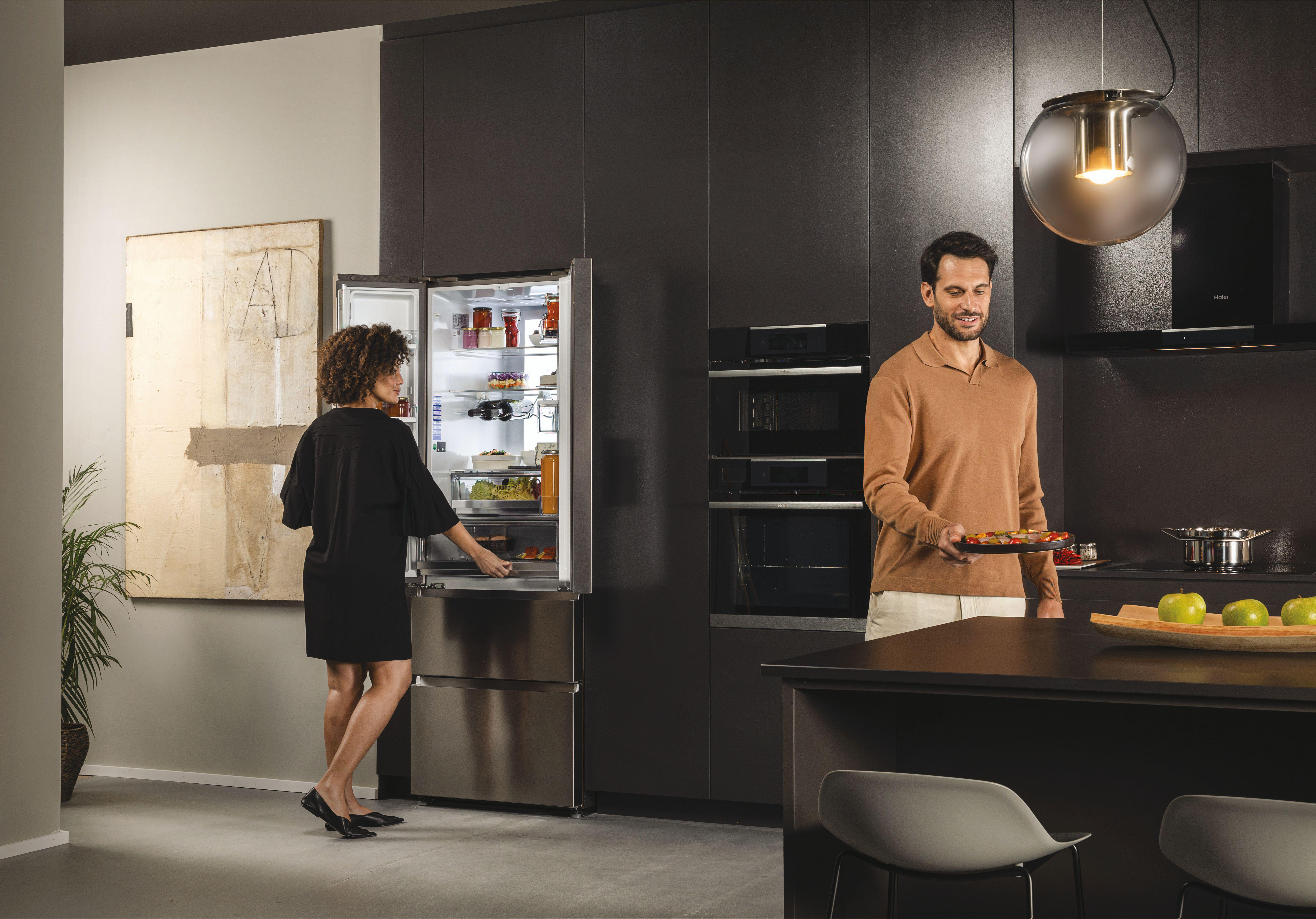 Frau entnimmt etwas aus einem Haier-Kühlschrank, während ein Mann Essen hält. Küche mit Geräten im Hintergrund.