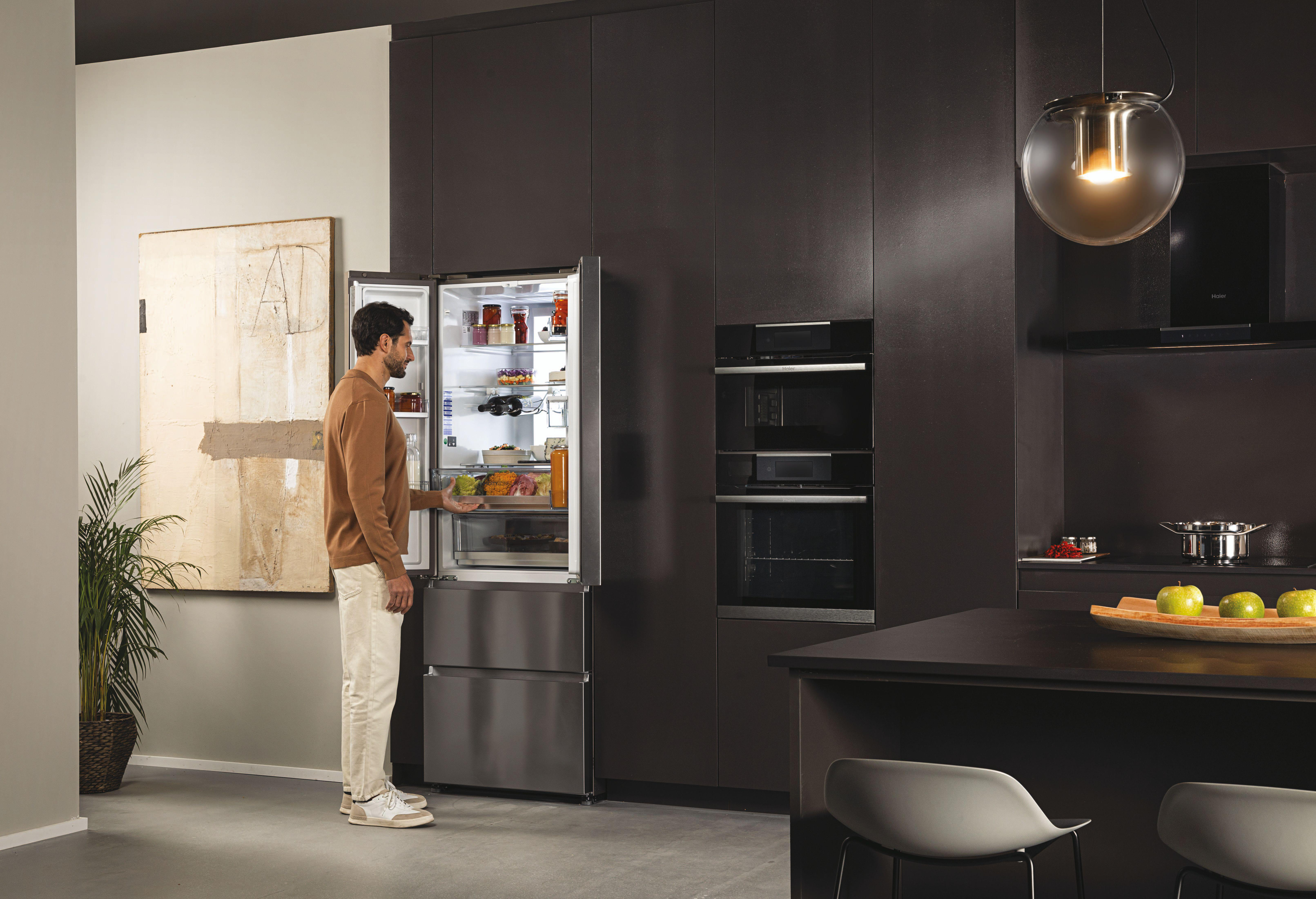 Mann nimmt Essen aus einem Haier-Kühlschrank. Eine andere Person hält Essen. Küche mit modernen Geräten im Hintergrund.