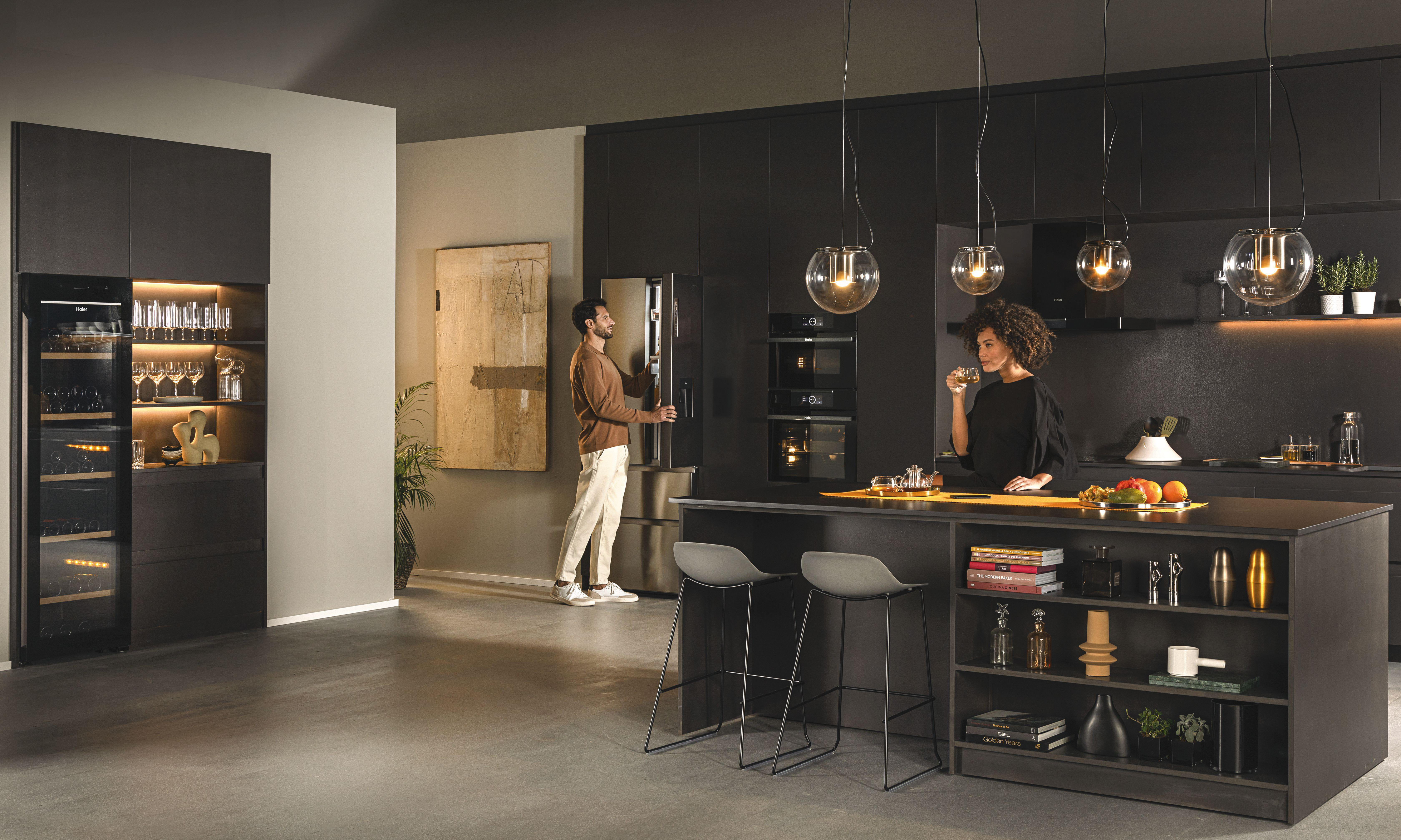 Moderne Küche mit dunklen Schränken, zwei Personen, einem Weinkühler und Glas-Pendelleuchten. Neutrales Farbschema.