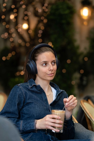 Frau mit Kopfhörern, die einen Kaffee hält. Sie ist in einem Café, und im Hintergrund sind Lichter.