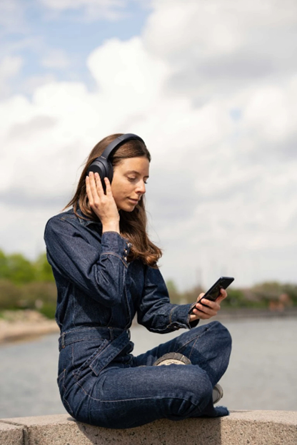 Frau in Jeansoverall sitzt und hört Musik über Kopfhörer, hält ein Handy.
