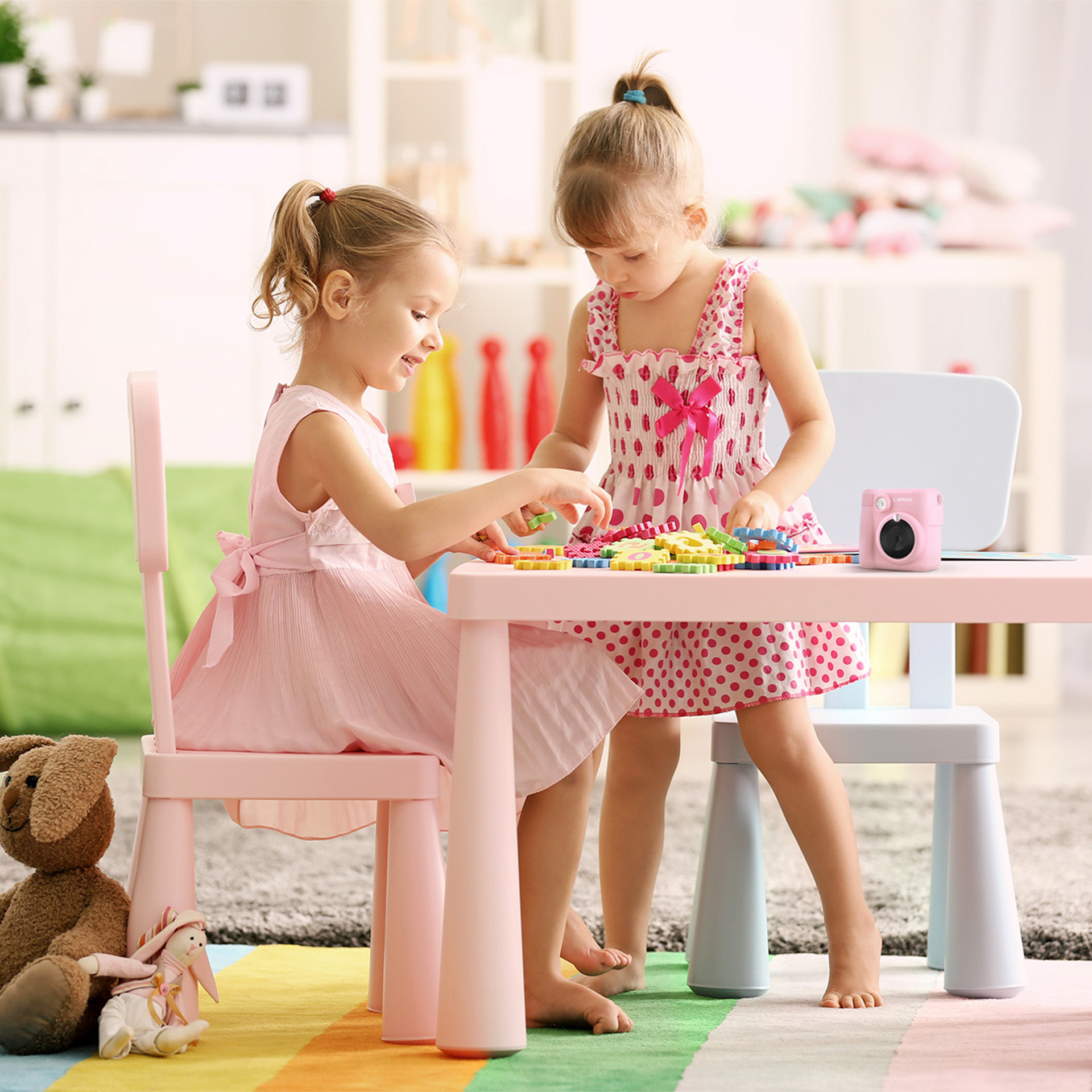 Két lány játszik egy rózsaszín asztalnál színes játékokkal. Maci és fényképezőgép.