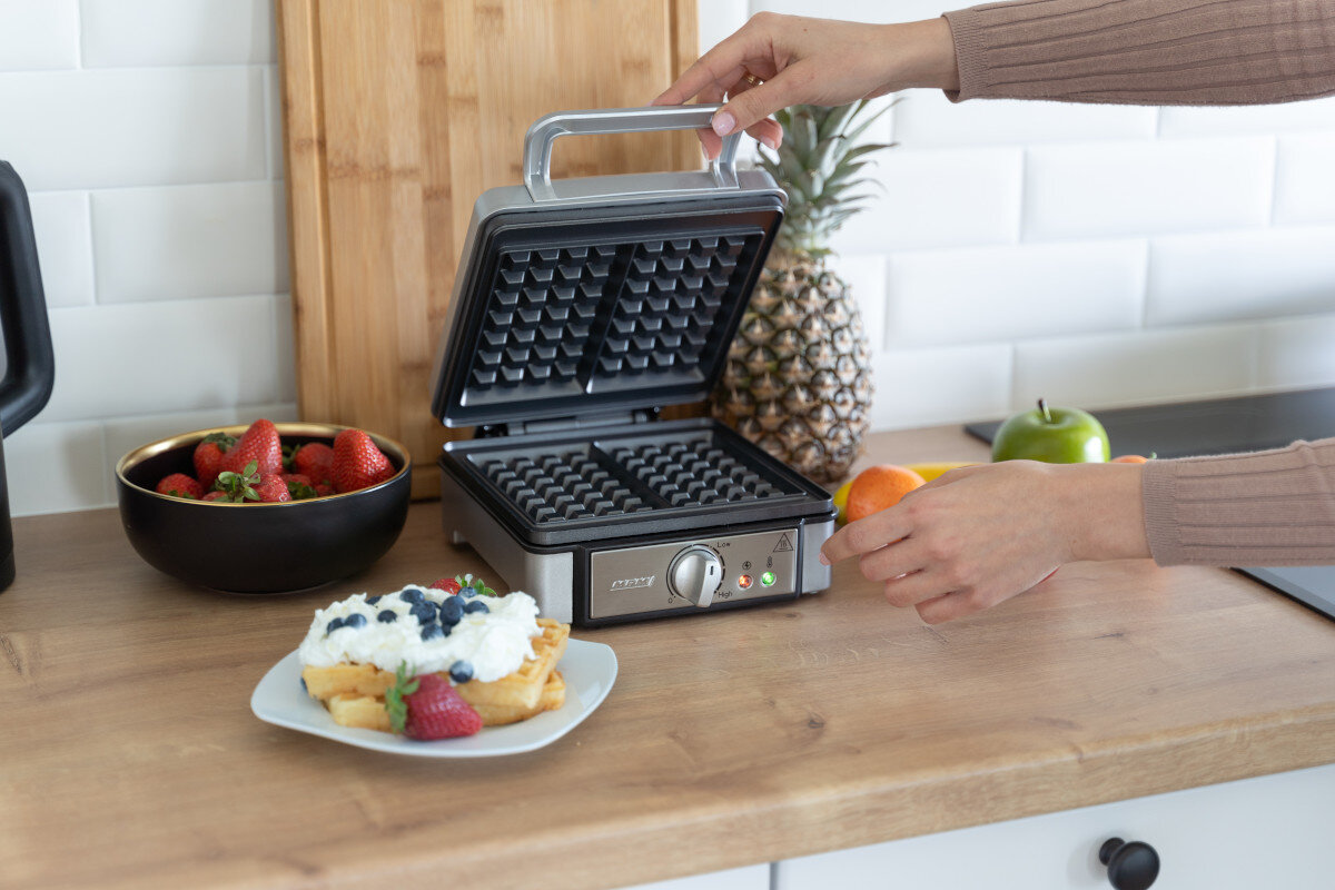 Osoba używa gofrownicy. Gofry z borówkami i kremem są na talerzu.