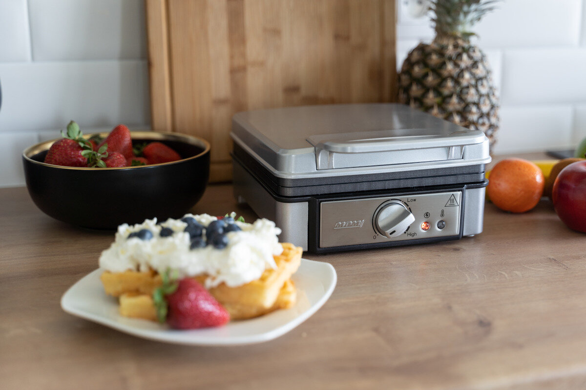 Gofr i gofrownica z goframi, truskawkami i bitą śmietaną na talerzu.