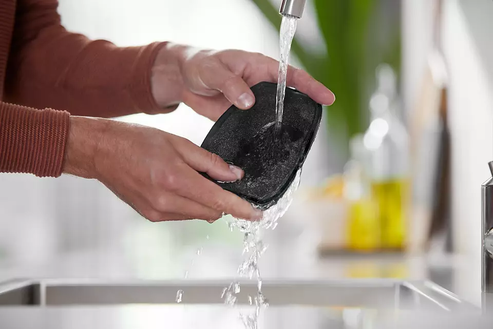 Ręce osoby myjącej czarny przedmiot pod kranem w otoczeniu kuchni.