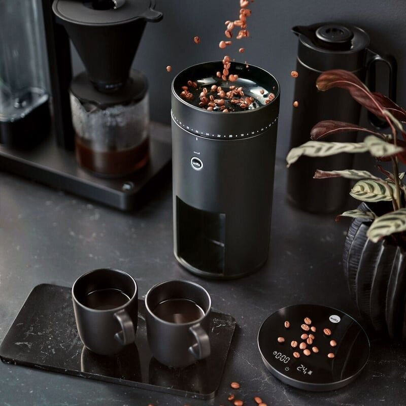 Czarny młynek do kawy z wpadającymi ziarnami, czarną wagą i dwoma czarnymi filiżankami.