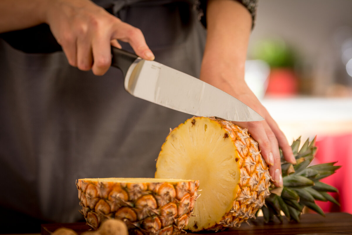Osoba kroi ananasa na pół dużym nożem na drewnianej desce do krojenia.