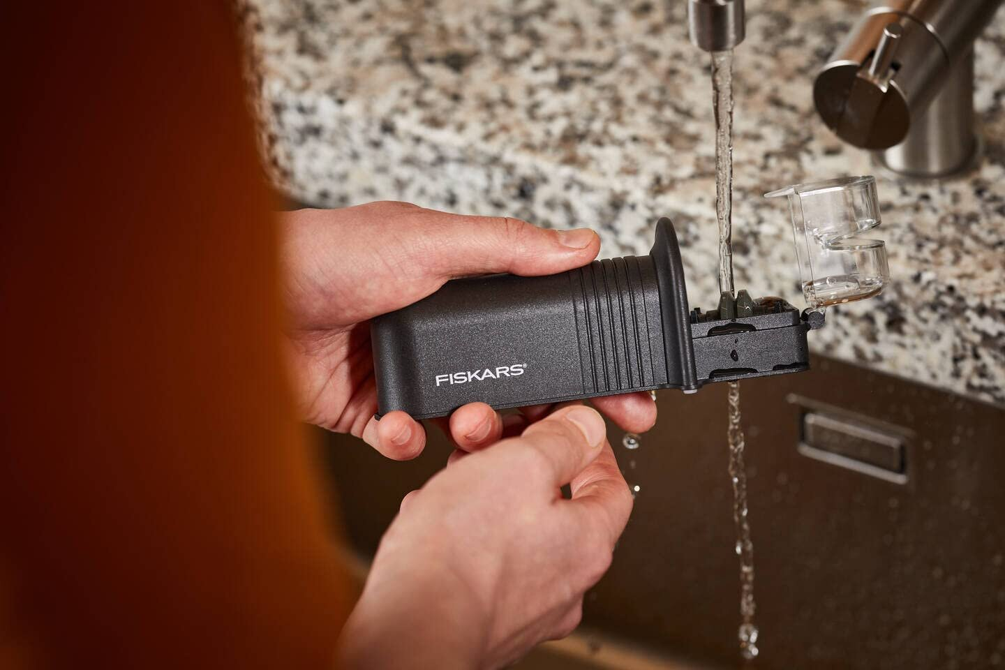 Dłonie myjące czarne narzędzie Fiskars pod bieżącą wodą w zlewie.