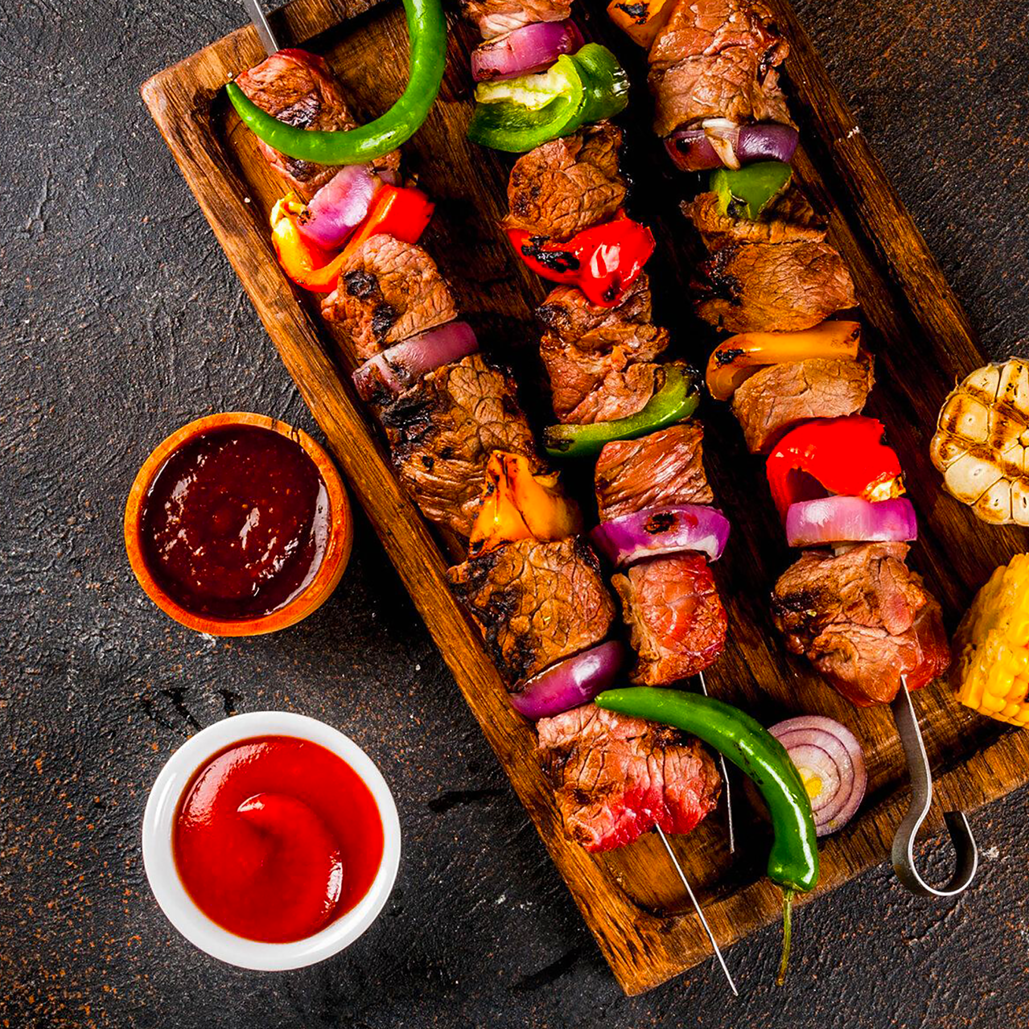 Grillezett hús és zöldségek nyárson, mártásokkal tálalva fa tálcán.