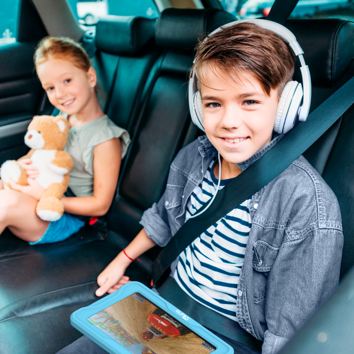 Children in car, boy with headphones, girl with teddy, looking at camera.