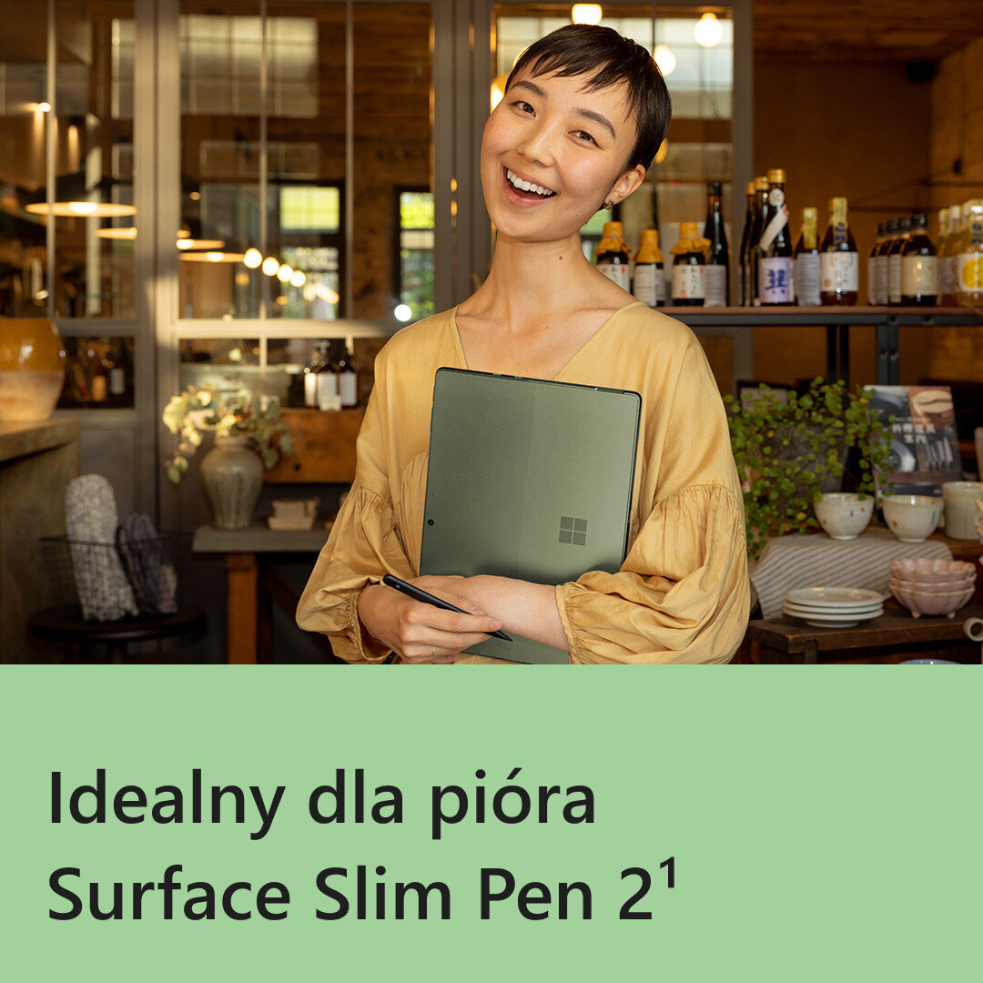 Kobieta w żółtej koszuli trzyma Surface Slim Pen i zielony laptop Surface, uśmiechając się do widza.