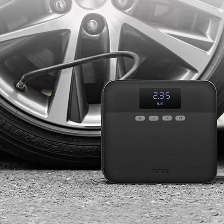 Czarna opona samochodowa z przenośną czarną sprężarką powietrza obok, wyświetlającą ciśnienie powietrza w barach.