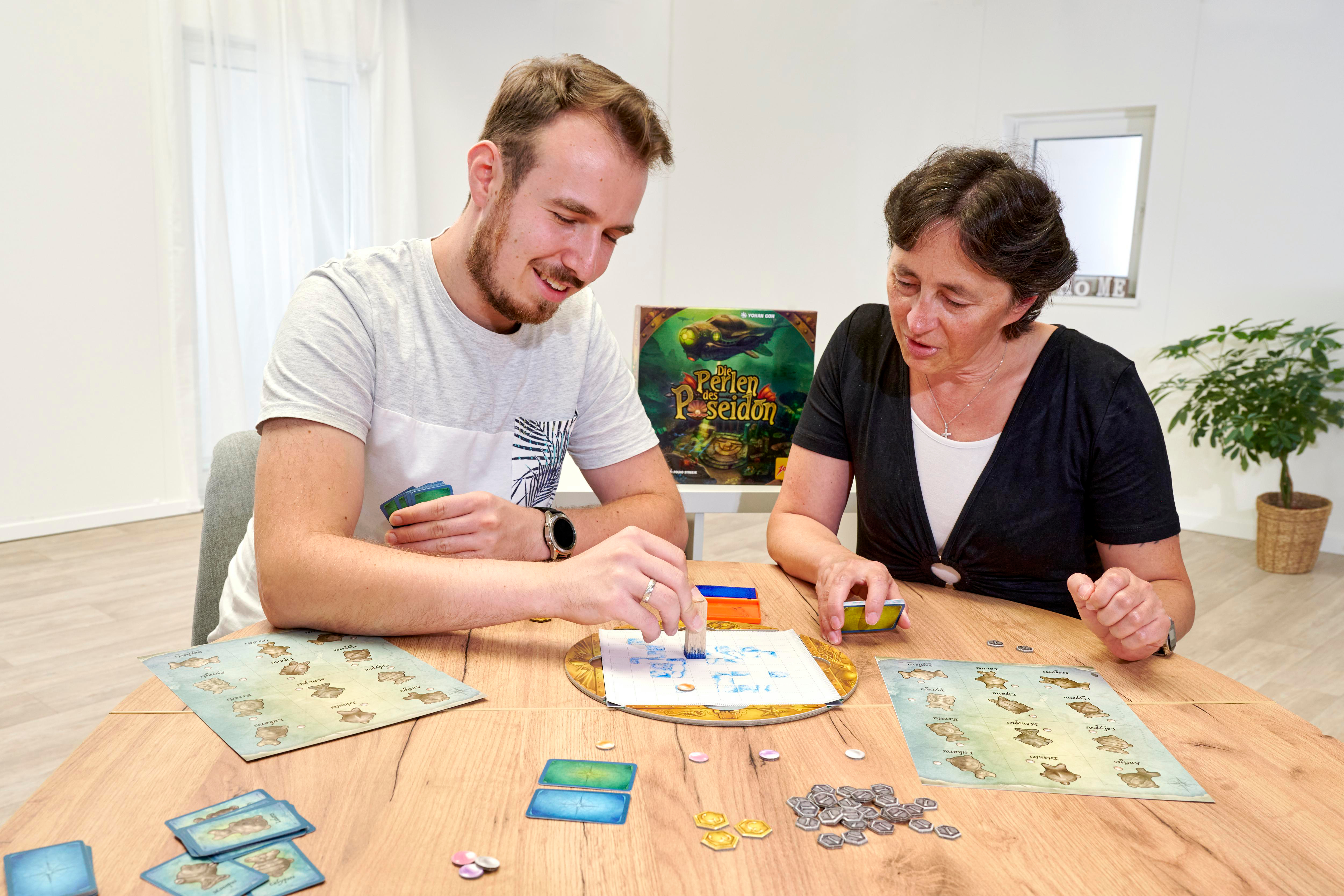 Zwei Personen spielen ein Brettspiel, mit Spielsteinen, Karten und einer Spielbox sichtbar.