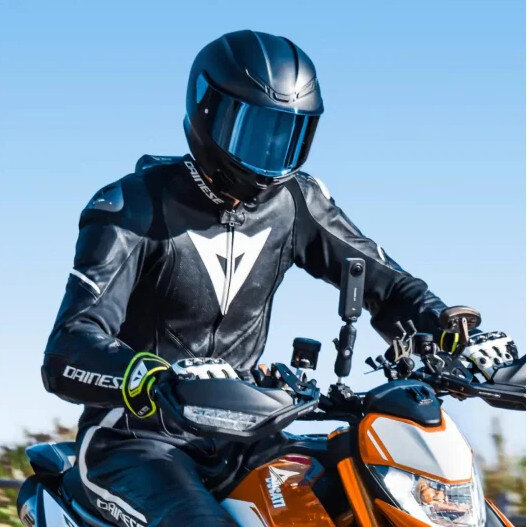 Motocyklista w czarnym stroju Dainese jedzie pomarańczowym motocyklem w słoneczny dzień.