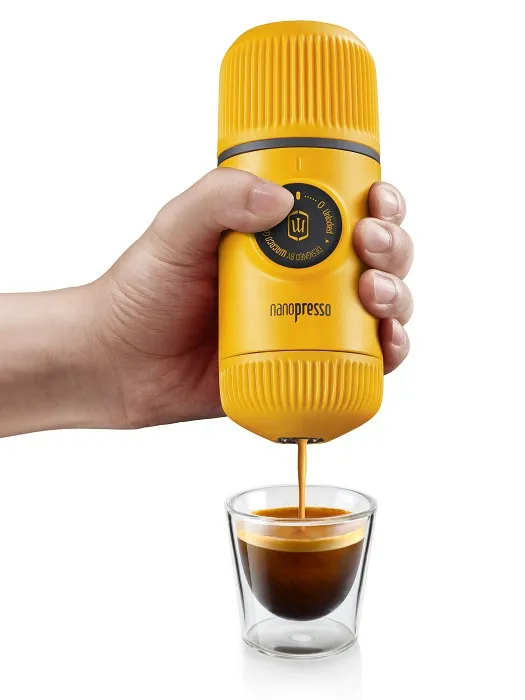 Egy kéz tart egy sárga hordozható eszpresszó gépet, kávét öntve egy pohárba.
