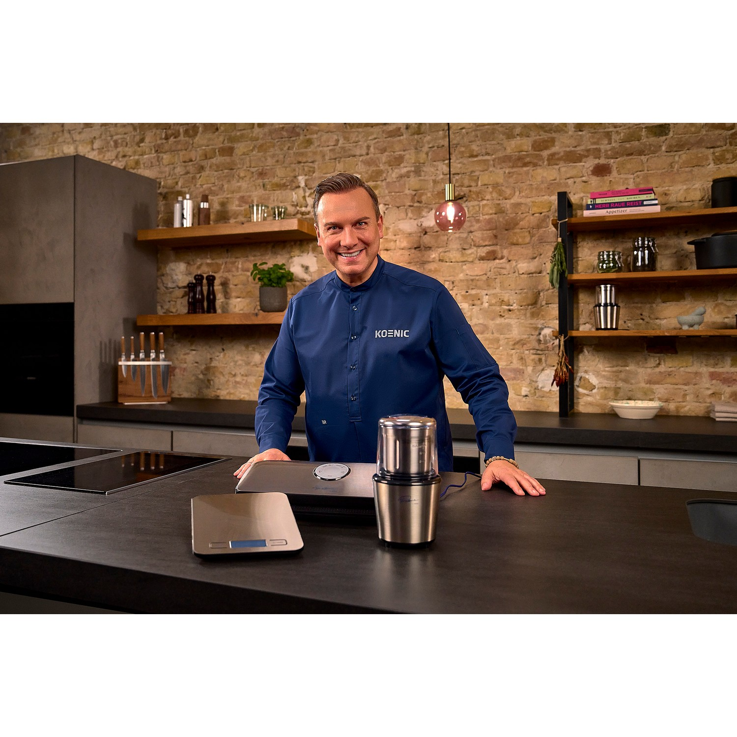 Een lachende man in een blauw overhemd staat achter een zwart aanrecht met een koffiezetapparaat.