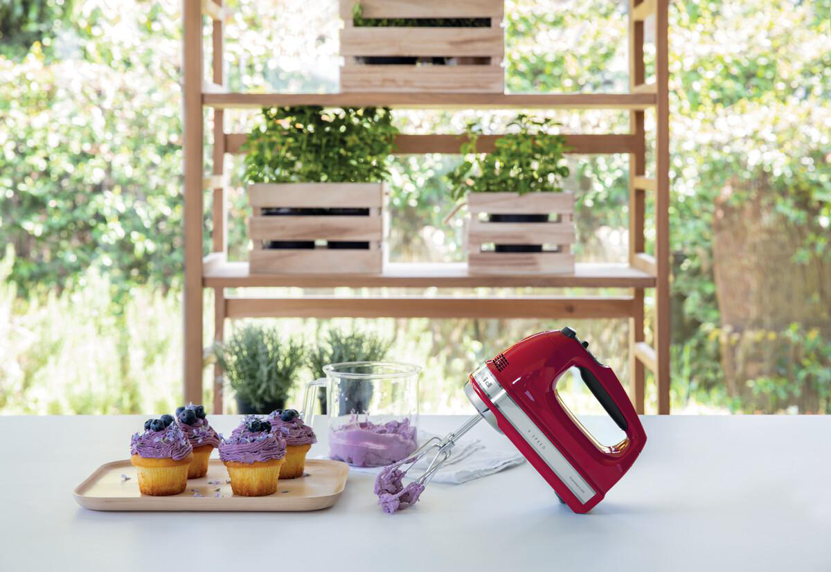 Roter Handmixer auf einem weißen Tisch mit Cupcakes und Pflanzen im Hintergrund.