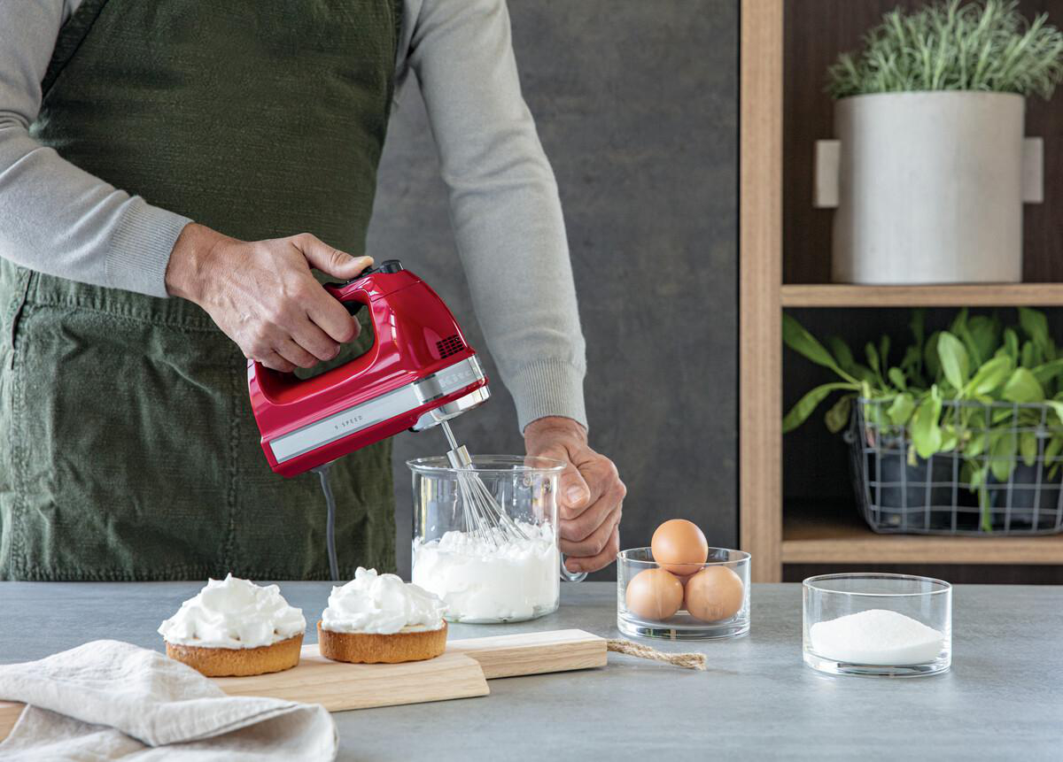 Eine Person, die einen roten Handmixer benutzt, um Sahne in einem Glas zu schlagen, mit Eiern und Kuchen auf dem Tisch.