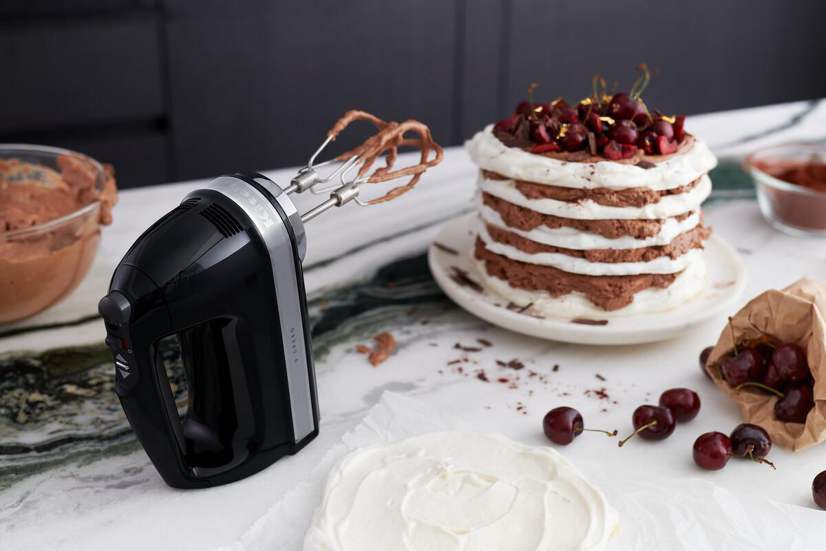 Schwarzer Handmixer mit Schlagbesen, neben Kuchen und Zutaten auf Marmoroberfläche.