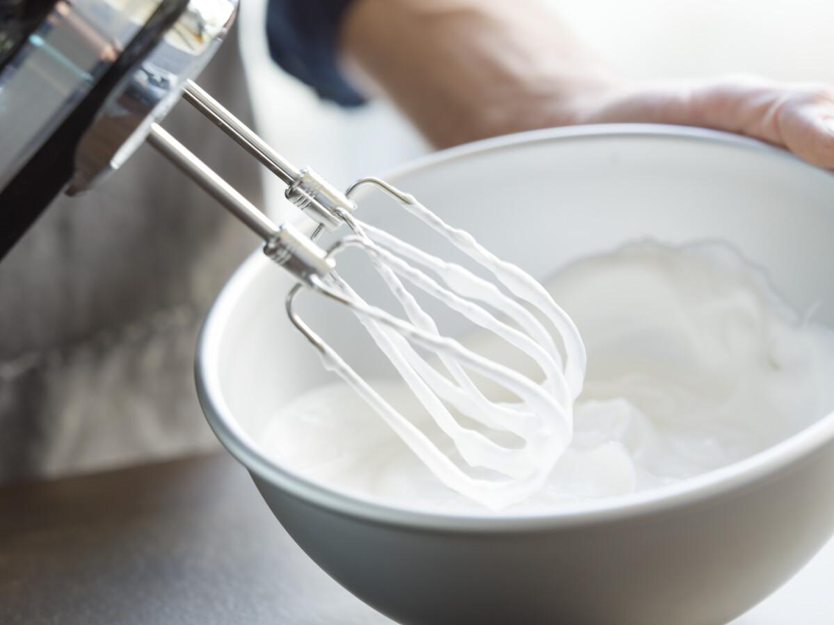 Handmixer mit Schneebesen in einer weißen Schüssel mit weißer Flüssigkeit. Hände halten den Mixer.