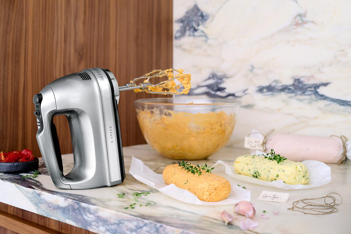 Silberner Handmixer mit Butter, Kräutern und anderen Zutaten auf Marmoroberfläche.