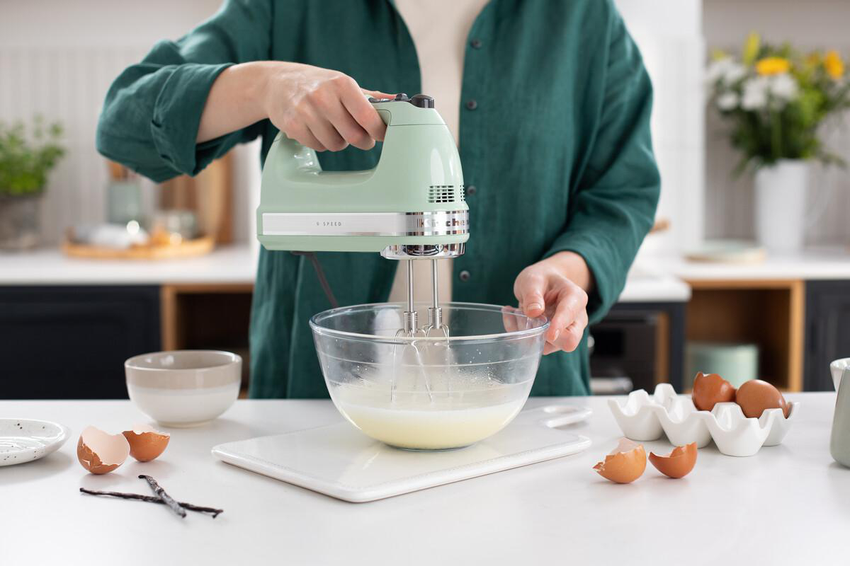 Person verrührt Zutaten in einer Glasschüssel mit einem grünen Handmixer, Eier und Schalen daneben.